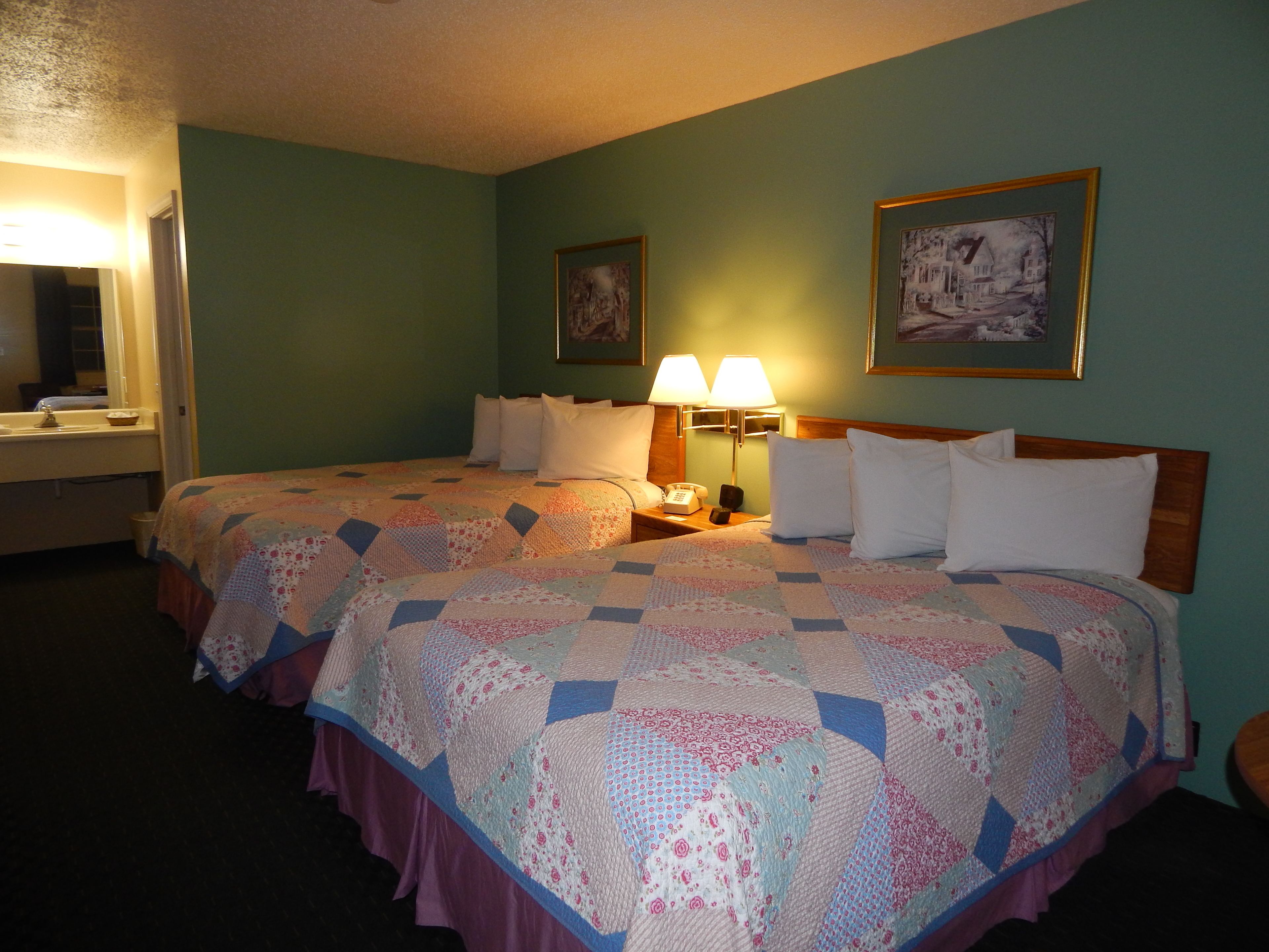A hotel room with two beds, a nightstand with a lamp, a painting on the wall, and a bathroom with a sink and mirror.