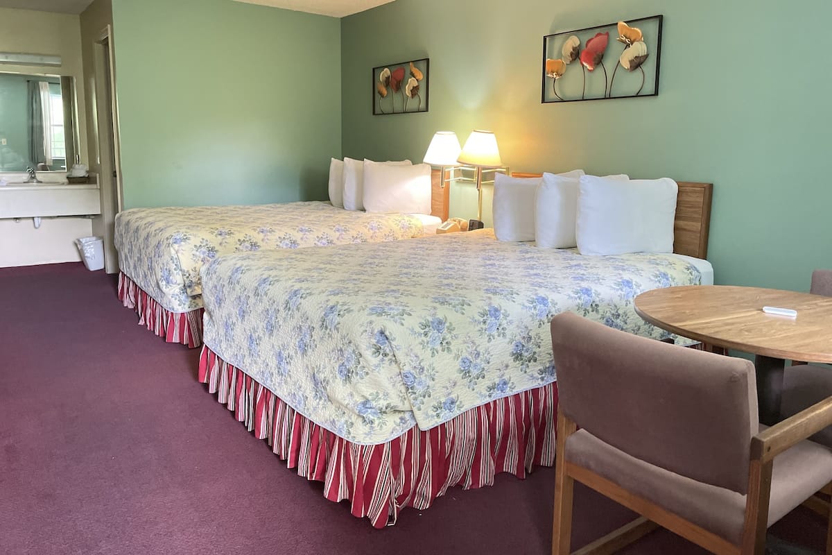 A hotel room with two beds, a round wooden table, a chair, a mirror, and two wall-mounted lamps.