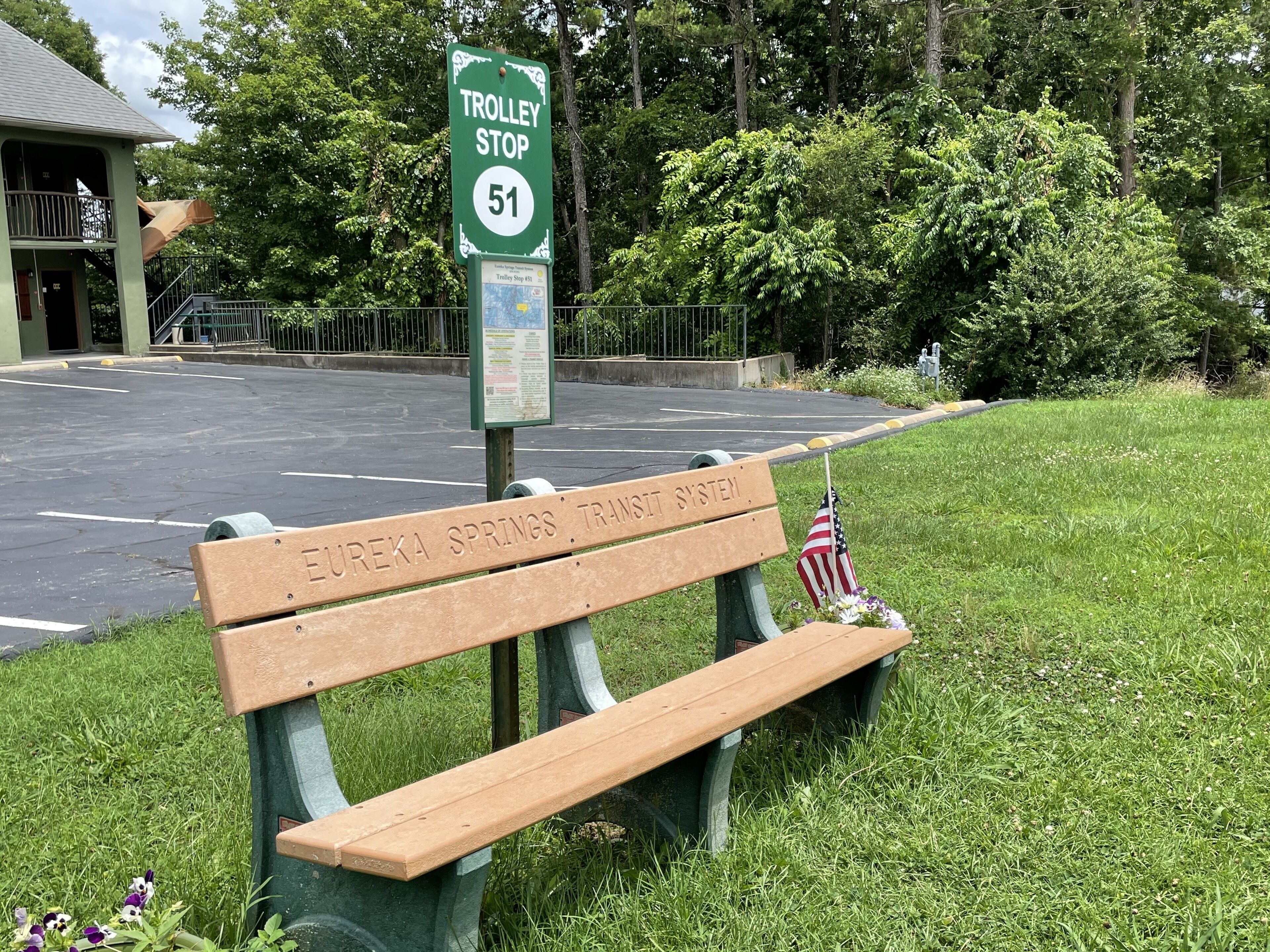 A trolley stop sign with a bench labeled "Eureka Springs' Transit System" and an American flag.