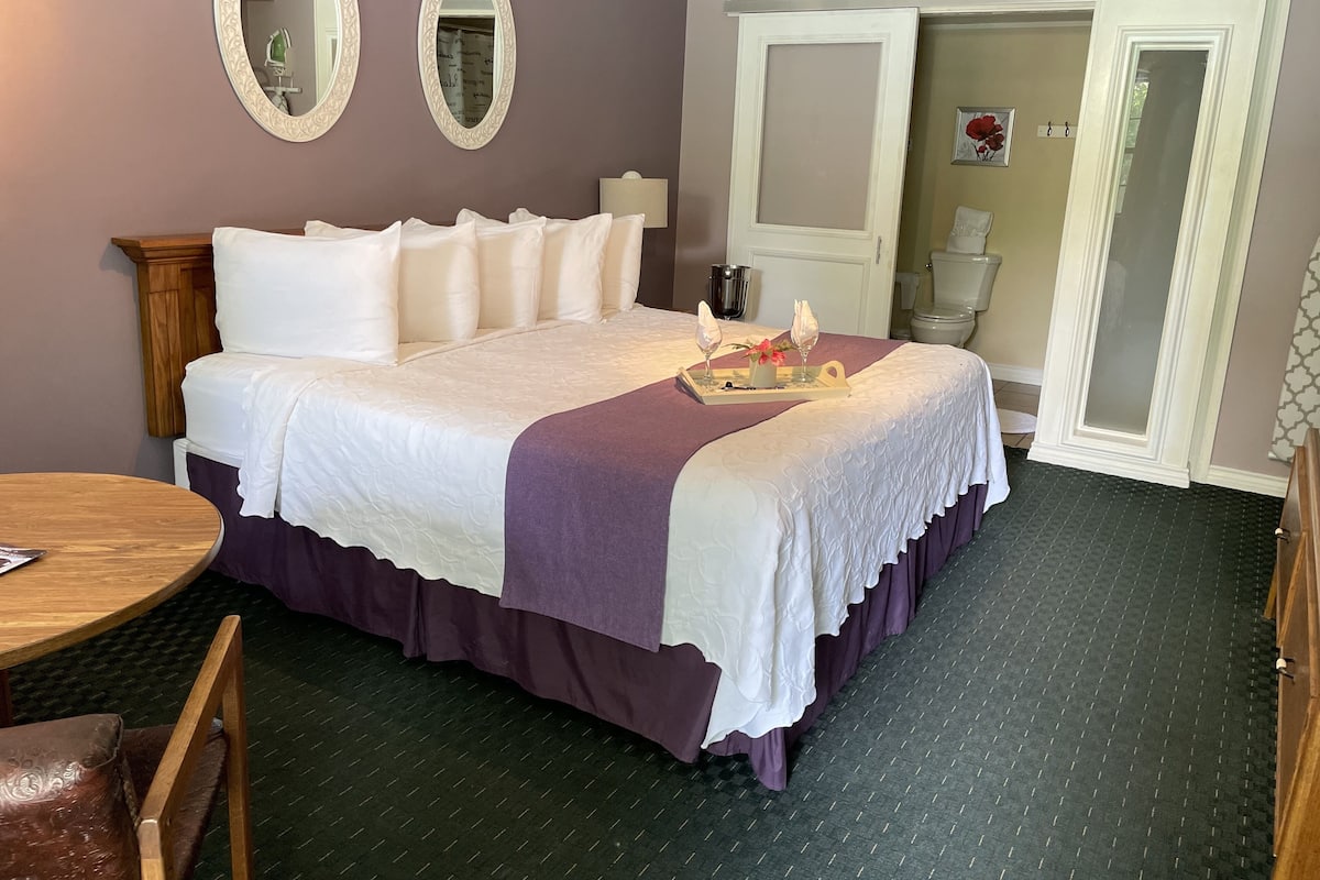 A neatly made bed with a purple bedspread, two wall mirrors, a wooden nightstand, a small round table, and a chair.