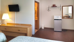 A hotel room with a wooden dresser, a bed, a television mounted on the wall, a microwave, and a small shelf with a cup and a candle. - Alpine Motel (Wisconsin Dells)
