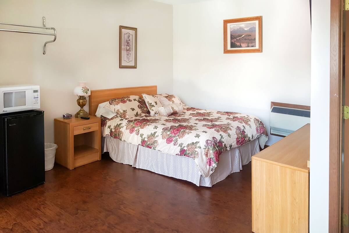 Un dormitorio con cama de madera, mesita de noche, un refrigerador pequeño, una lámpara y un cuadro en la pared.