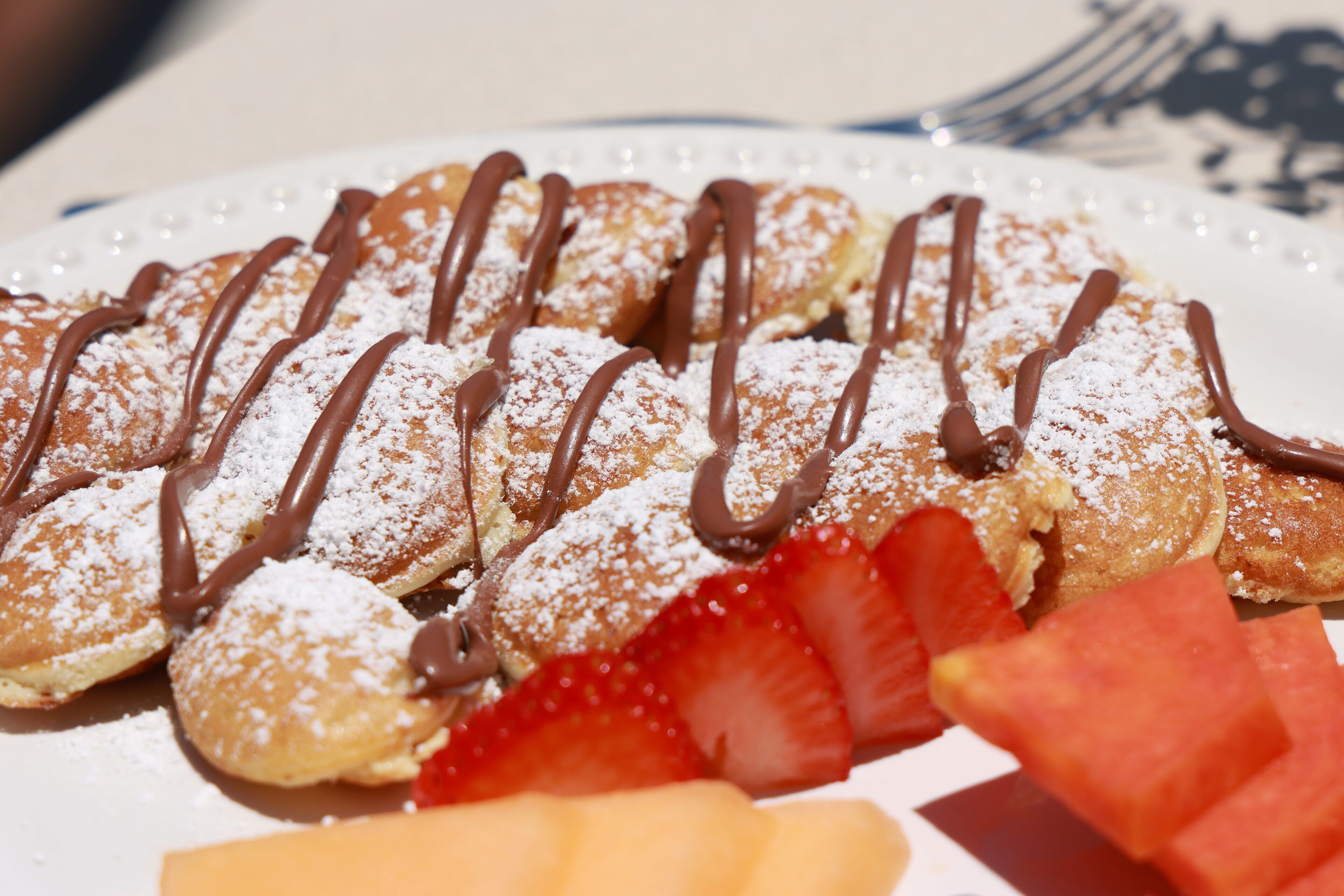 A plate of pastries with chocolate drizzle and powdered sugar, accompanied by fresh fruit slices.