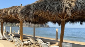 Una zona de playa con sombrillas de paja y sillones blancos.