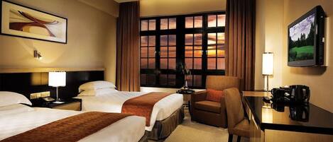 A hotel room with two beds, a desk, a chair, a TV, and a large window with curtains.