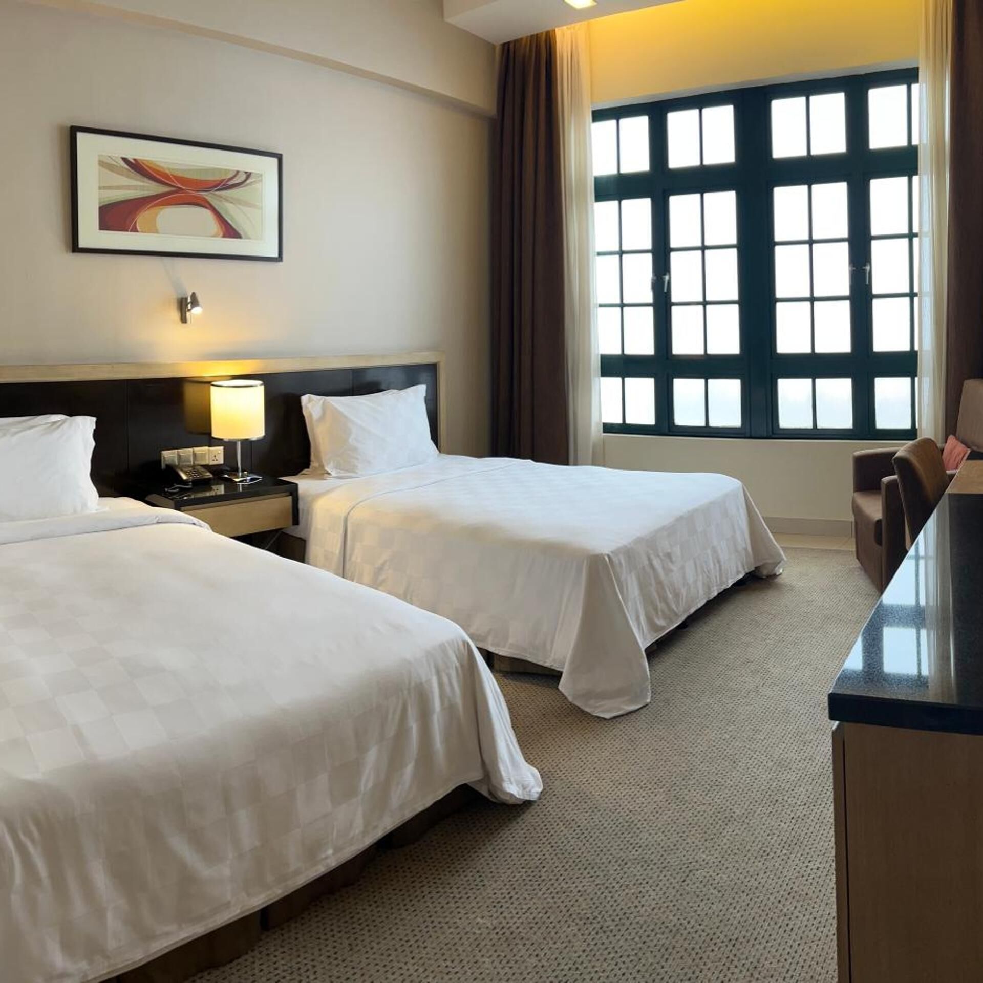 Une chambre d’hôtel avec deux lits, un bureau, une chaise, une table de chevet, une lampe, un tableau et une fenêtre avec des rideaux.