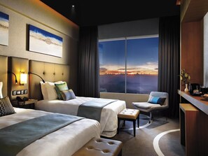 Habitación de hotel con dos camas, un escritorio, una silla y un amplio ventanal con vista al atardecer.