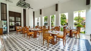 A spacious dining area with multiple tables and chairs, large windows offering views of the outdoors, and a patterned floor. - Inna Bali Heritage Hotel (Denpasar)