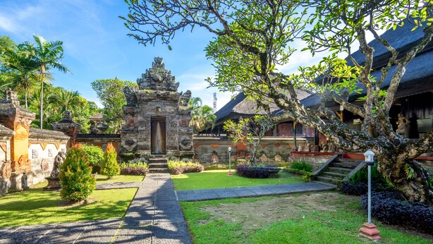 A courtyard with a stone pathway leading to a temple-like structure, surrounded by lush greenery and traditional architecture. - Inna Bali Heritage Hotel (Denpasar)