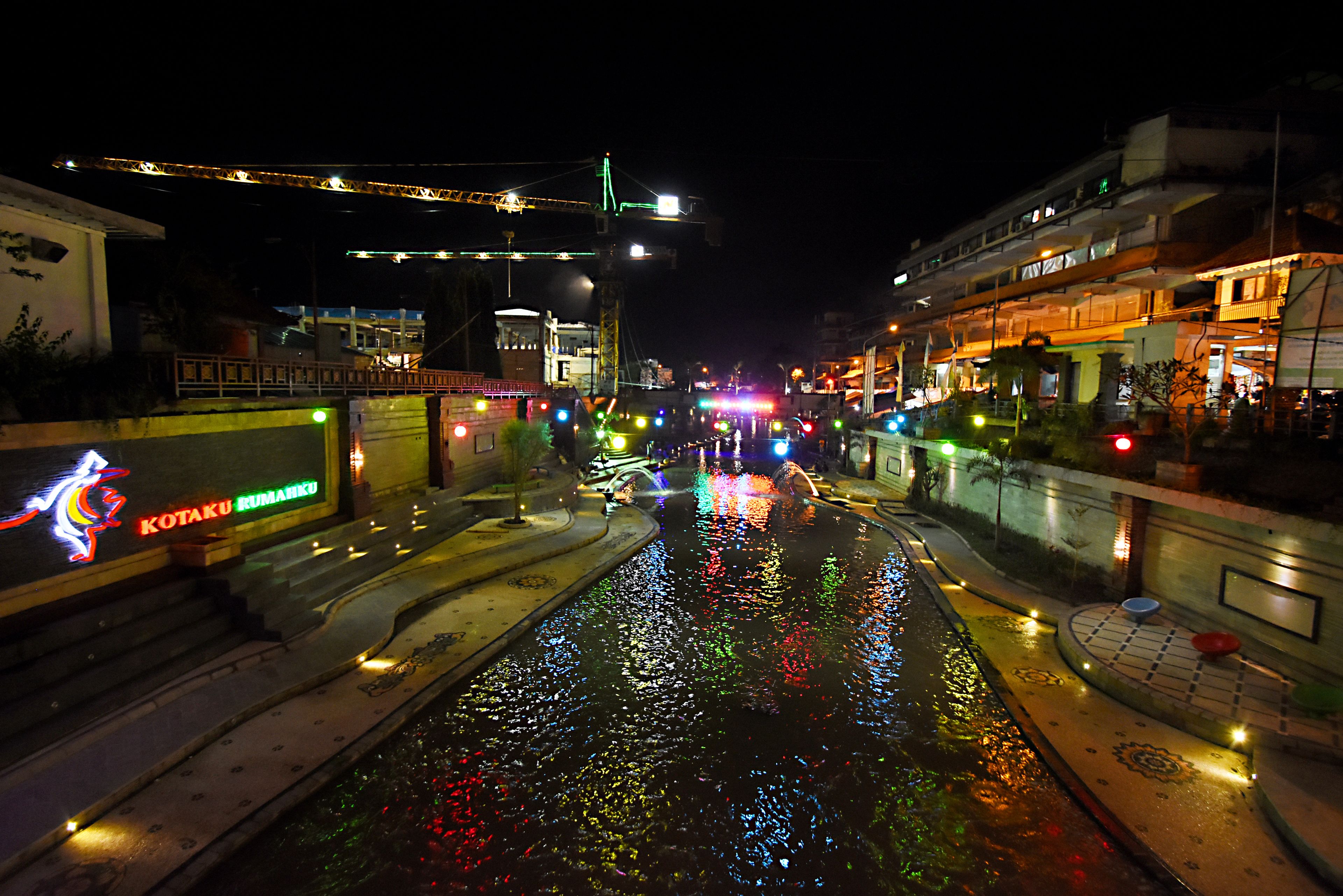 Una escena nocturna en la calle con un canal que refleja luces de colores, un puente y edificios con letreros iluminados.