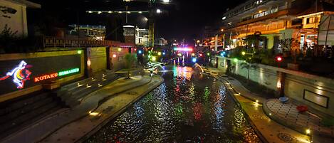 Una escena nocturna en la calle con un canal que refleja luces de colores, un puente y edificios con letreros iluminados.