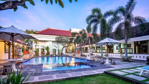 A swimming pool surrounded by outdoor seating, palm trees, and a building with a red roof. - Inna Bali Heritage Hotel (Denpasar)