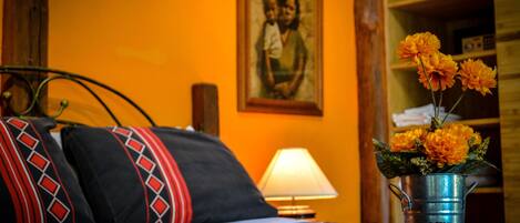 A bed with a patterned bedspread, a metal bucket with flowers, a lamp, and a framed picture on the wall.