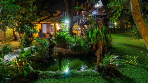 Un jardín bien iluminado con un estanque, rodeado de abundante vegetación y una casa tradicional con una escalera que conduce a un balcón.