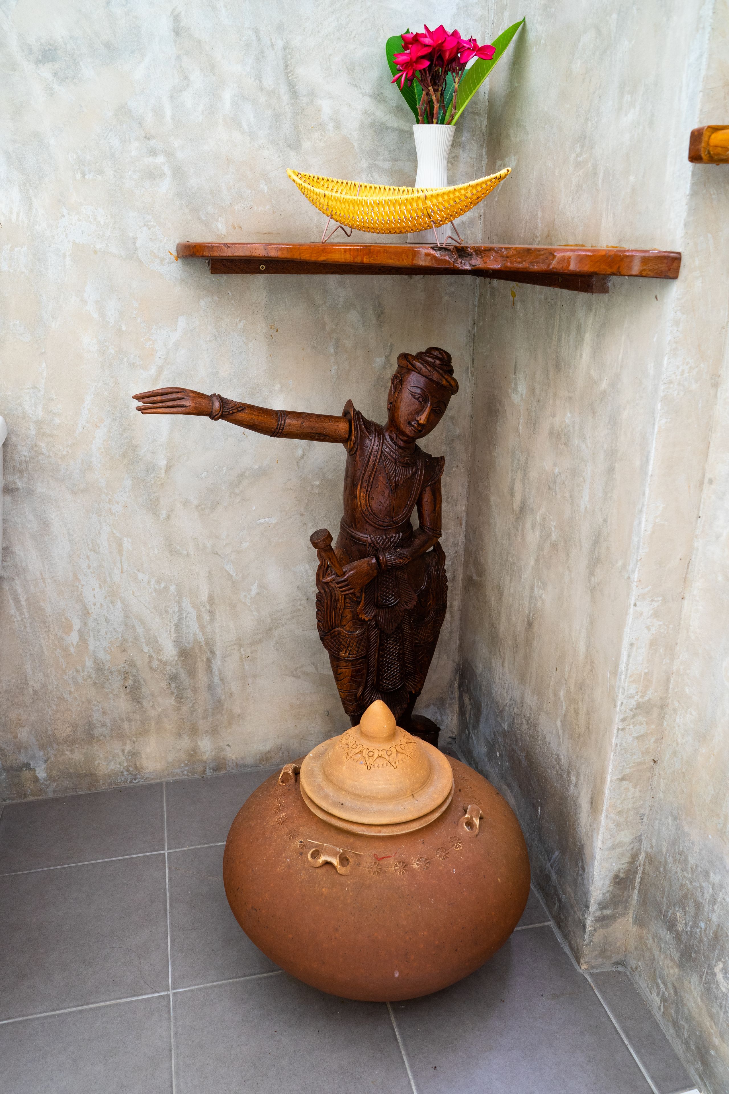 Una estatua de una figura con un brazo extendido, una cesta tejida y una olla de barro sobre una estantería frente a una pared con textura.