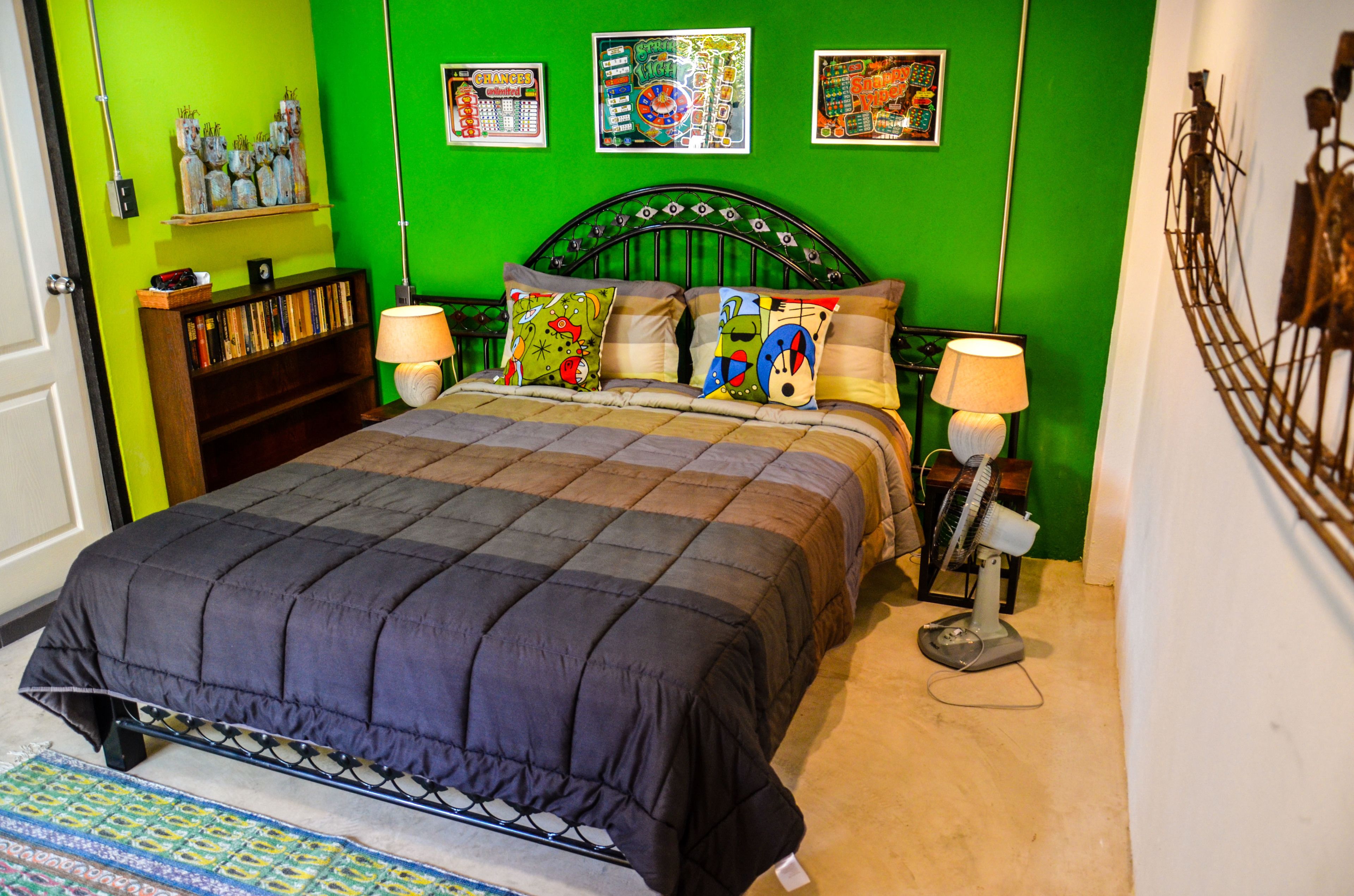 A bedroom with a green wall, a metal bed frame, a quilted bedspread, two bedside lamps, and a small bedside table with a lamp.