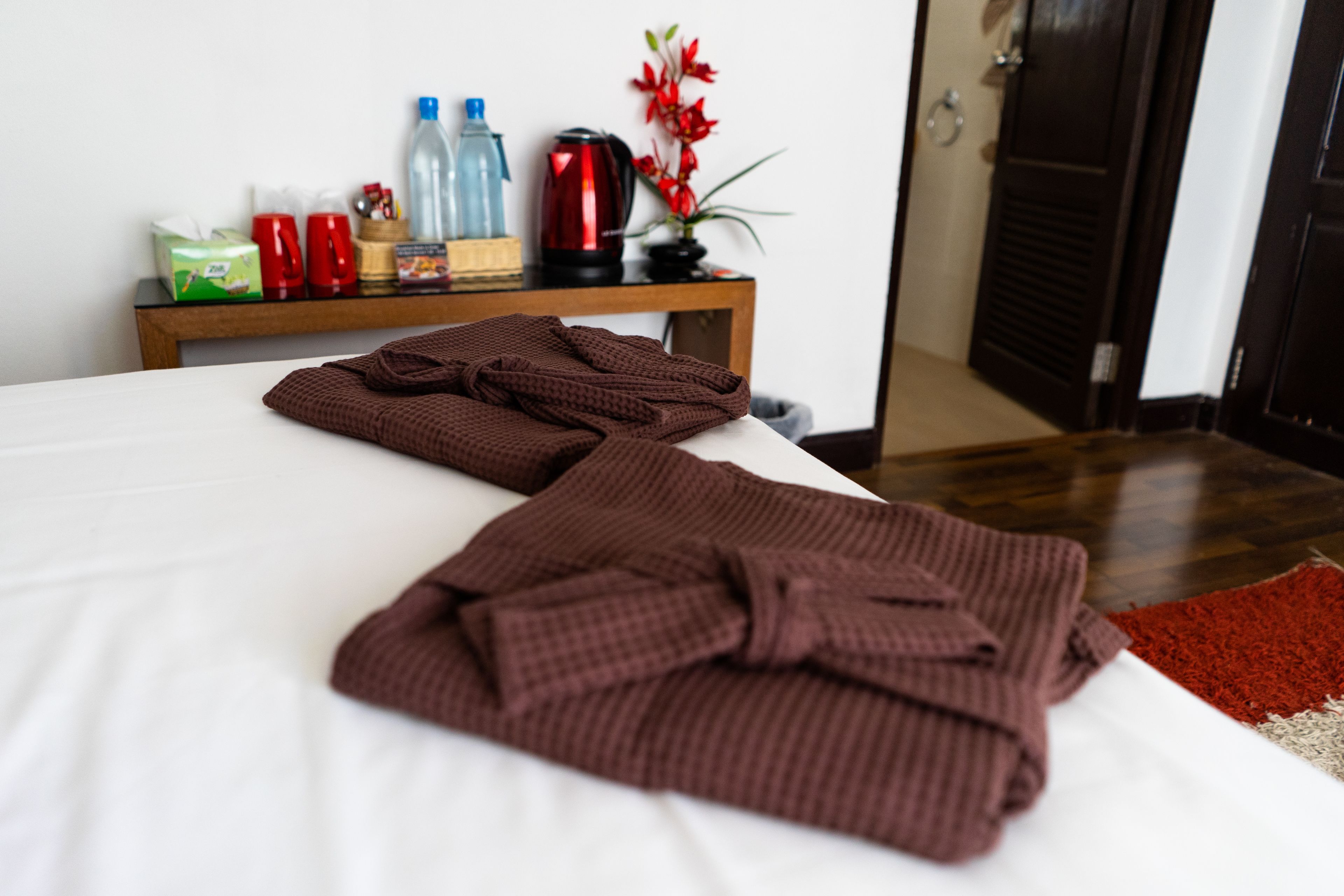 A neatly made bed with two folded brown blankets, a wooden nightstand with various items including a red kettle, bottles, and a small plant.