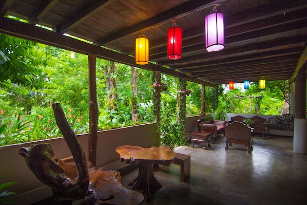 A covered outdoor area with wooden furniture, hanging lanterns, and lush greenery visible through the windows.