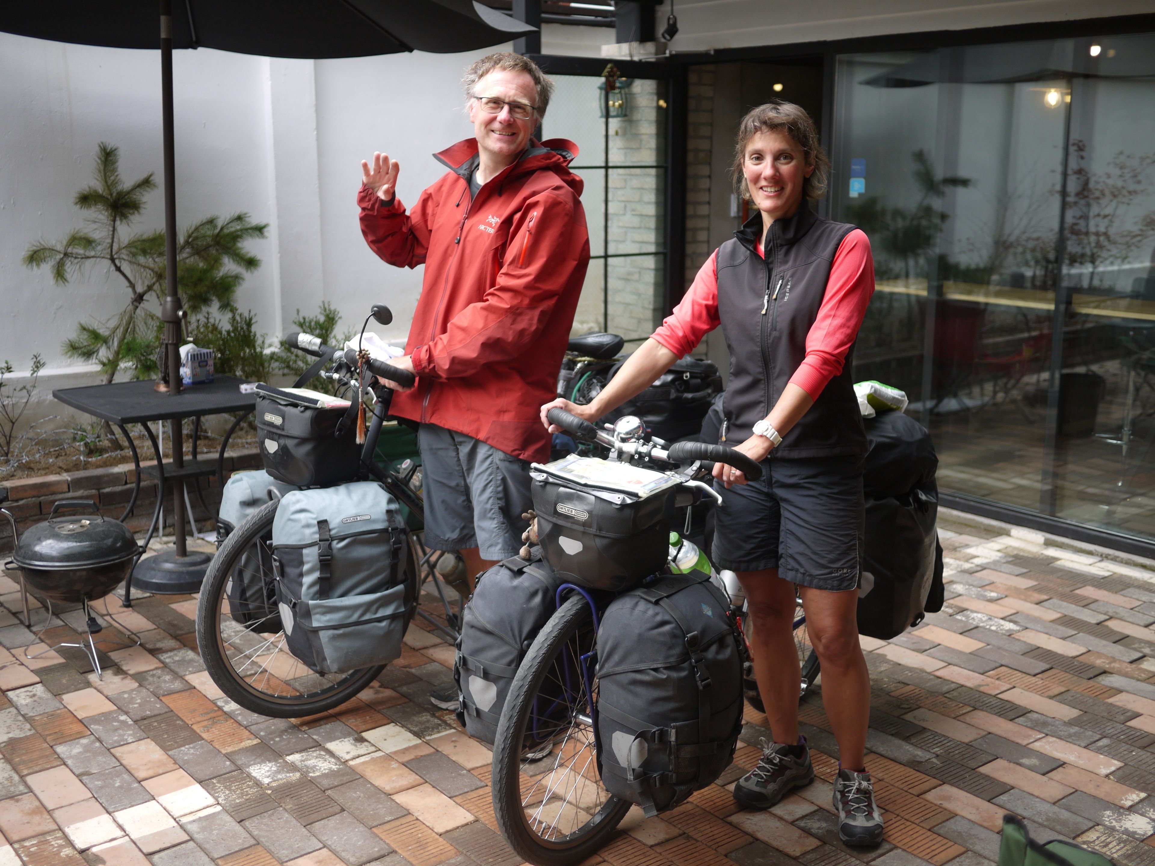Zwei Personen stehen neben einem Fahrrad mit mehreren Taschen, in einem Außenbereich mit Fliesenboden und einem Grilltisch.