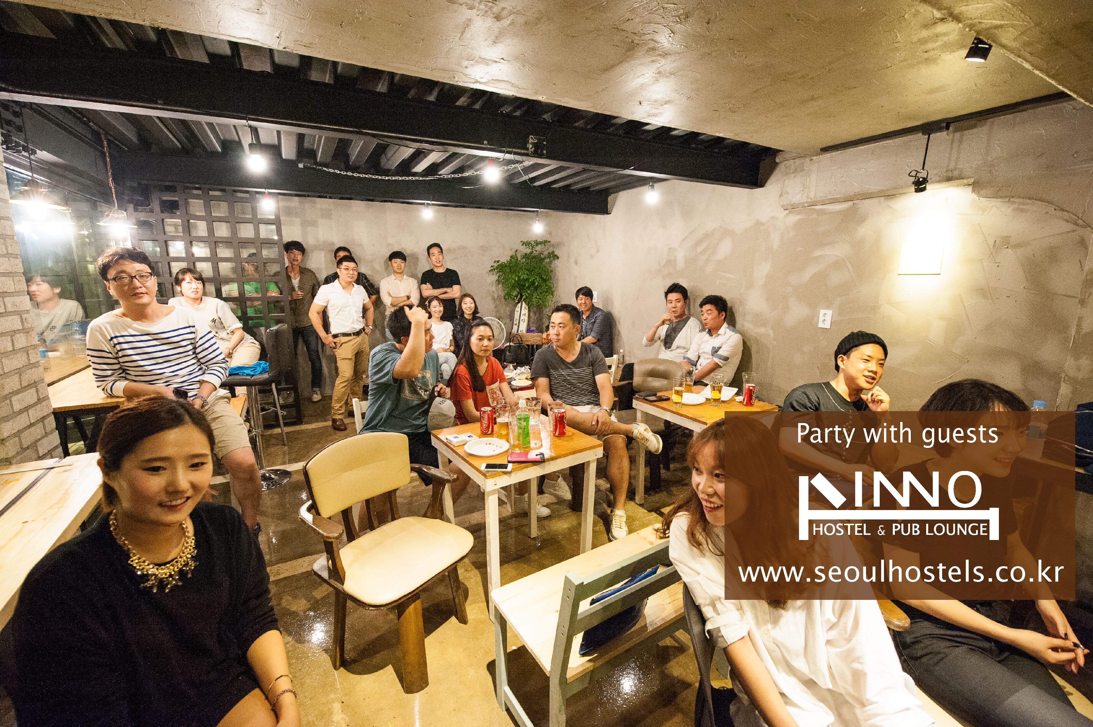A group of people in a hostel lounge with tables and chairs, a brick wall, and a hanging light fixture.