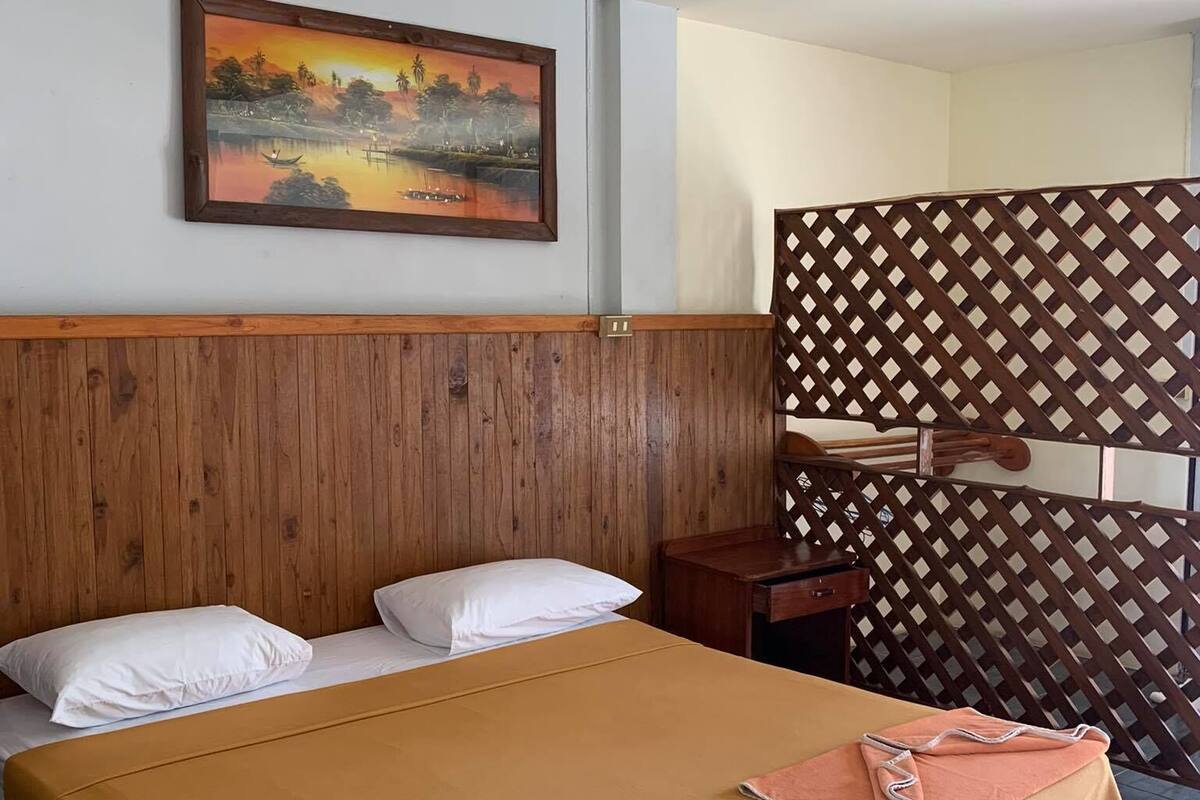 A hotel room with a wooden headboard, a large bed with a beige bedspread, two pillows, a wooden nightstand, a small table, and a decorative wooden lattice partition.