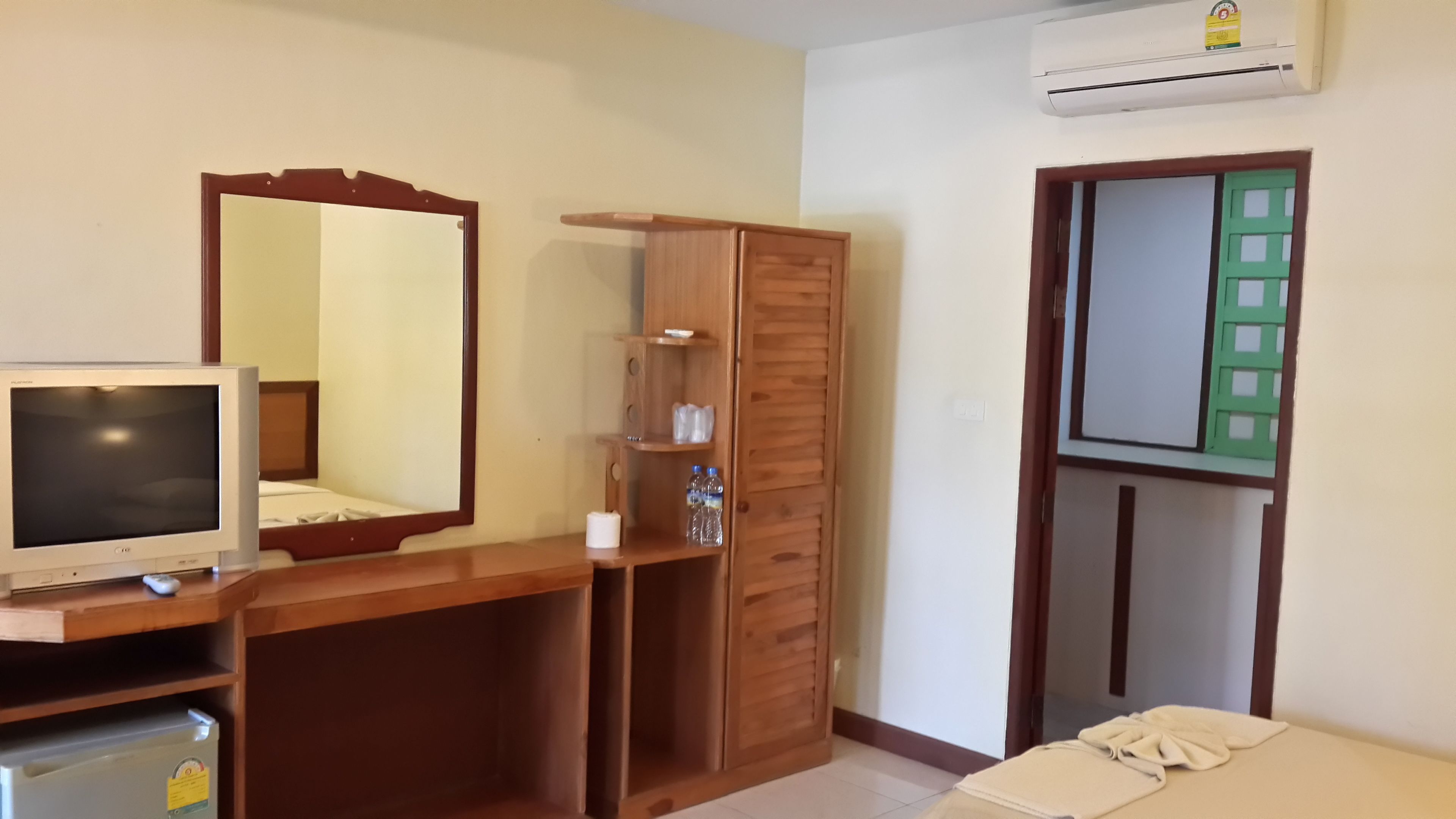 A bedroom with a wooden wardrobe, a mirror, a television, and a green-framed door.