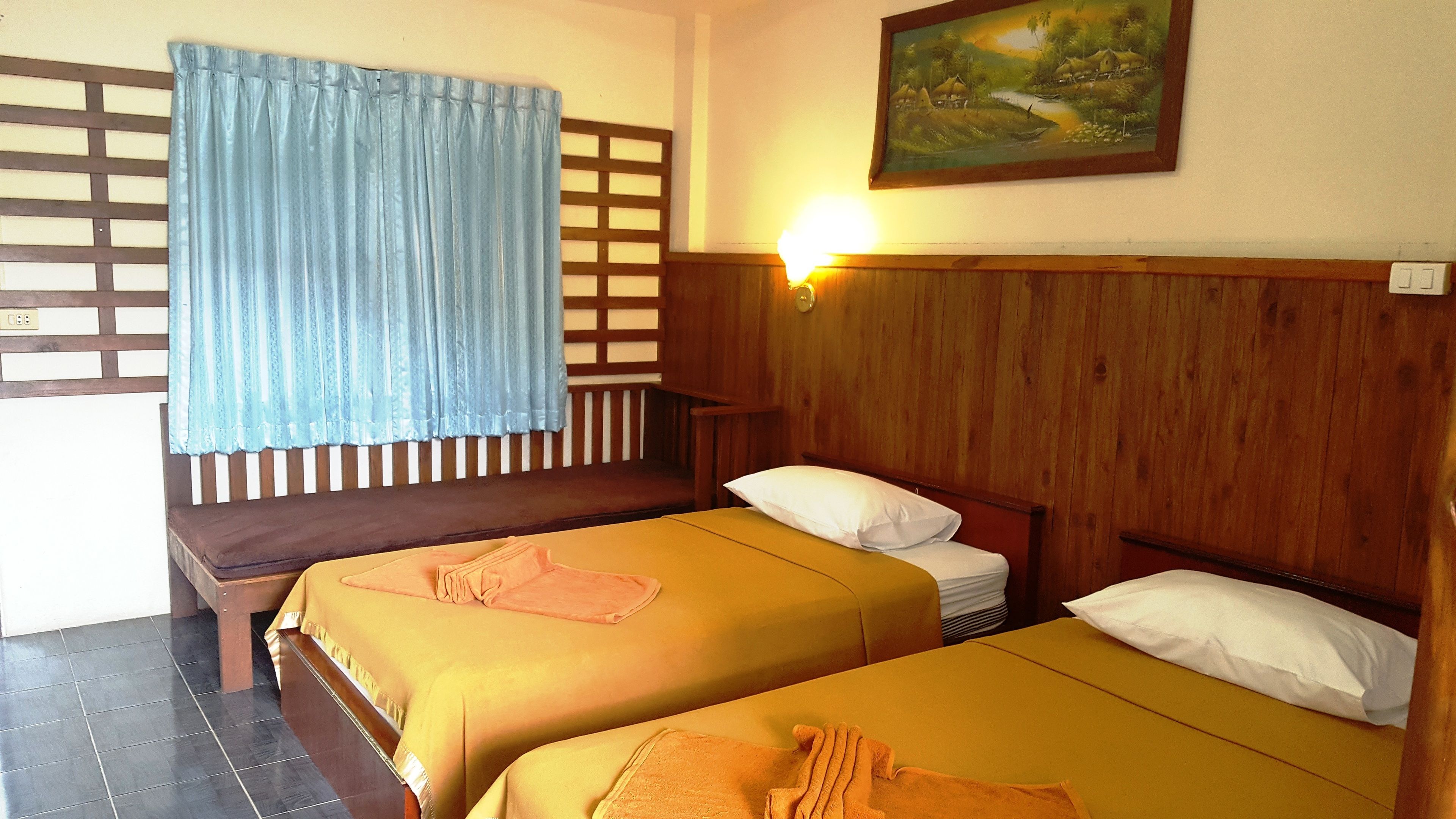 A room with two beds, a wooden headboard, a window with a blue curtain, a wall-mounted light, and a painting.