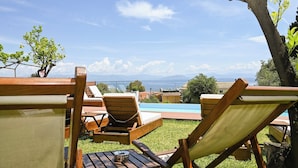 Zona junto a la piscina con sillas reclinables de madera y una mesa, con vista a un cuerpo de agua y montañas a lo lejos.