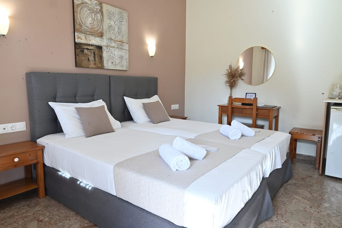 Una cama bien hecha con sábanas blancas y un cabecero gris, una mesita de noche con cajón, un espejo redondo en la pared y una pequeña mesa con un jarrón de plantas secas.