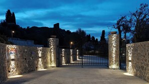 Un portón de piedra con una cerca de metal, iluminado por luces, que da a un camino.