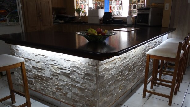 A kitchen with a stone backsplash, a central island with a fruit bowl, wooden bar stools, and a window with stained glass. - Sleepers Villa (Polokwane)