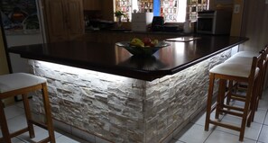 A kitchen with a stone backsplash, a central island with a fruit bowl, wooden bar stools, and a window with stained glass. - Sleepers Villa (Polokwane)