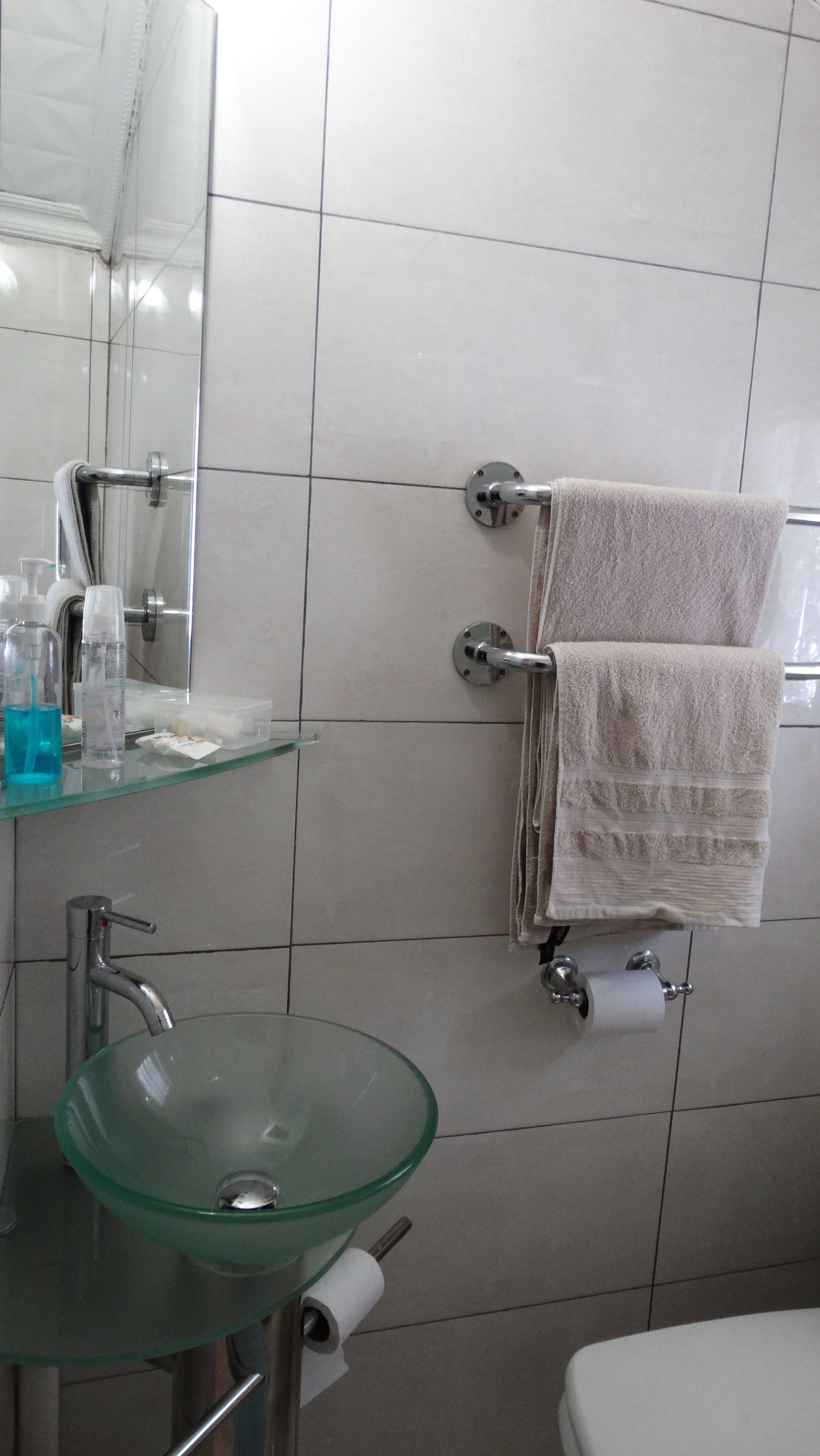 A bathroom with a glass sink, a mirror, a towel rack with two towels, a toilet paper holder, and a shelf with toiletries.
