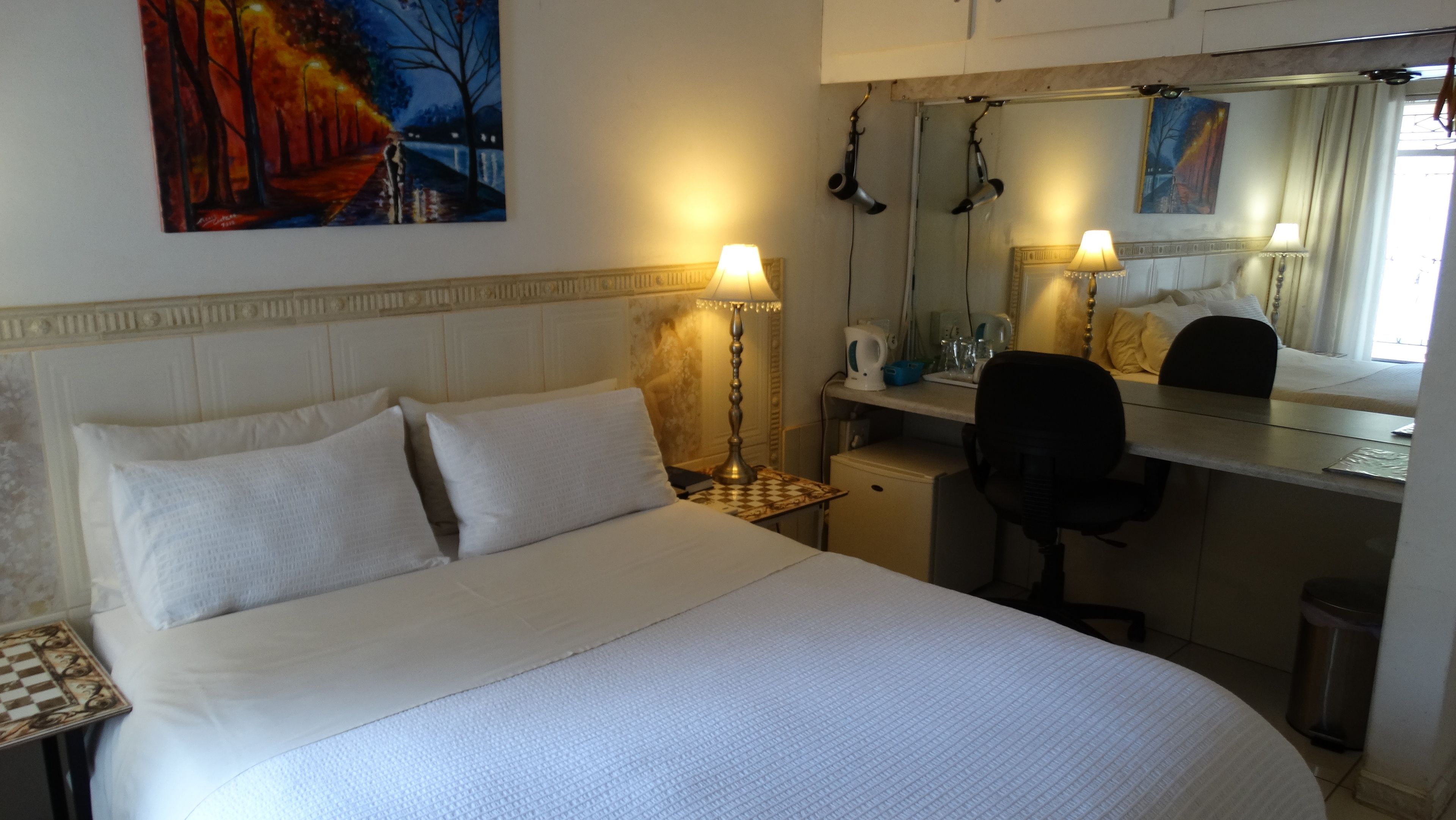 A hotel room with a large bed, two bedside lamps, a desk with a chair, a mirror, and a painting on the wall.