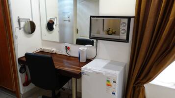 A small room with a wooden desk, a white refrigerator, a mirror, and a wall-mounted TV.