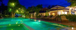 A nighttime swimming pool area with lounge chairs and umbrellas, surrounded by lush greenery and illuminated by ambient lighting.