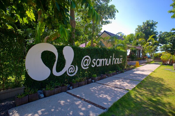 A landscaped area with a large sign reading "Samui Haus" surrounded by greenery and a pathway.