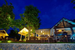 A well-lit outdoor dining area with tables and chairs, surrounded by trees and a stone wall.