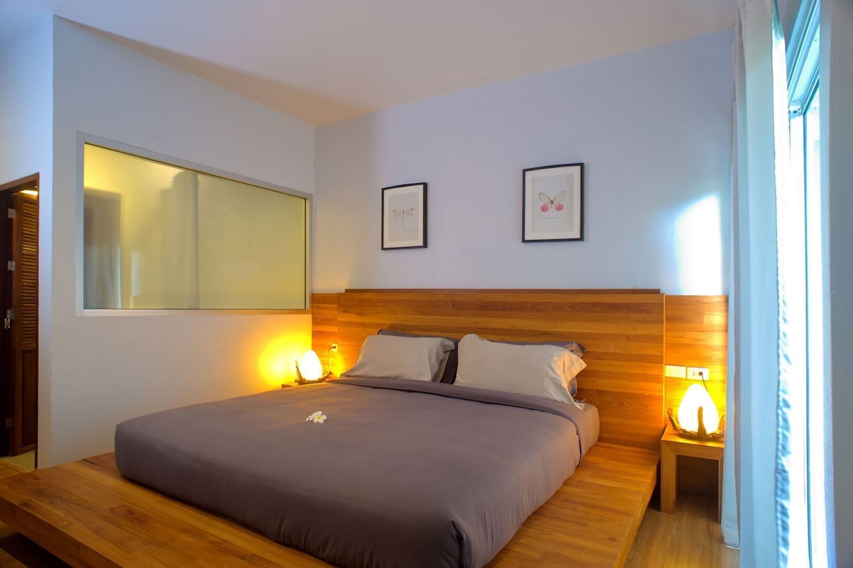 Quarto com uma cama, duas luminárias de cabeceira, uma mesinha de cabeceira, um espelho grande e duas fotografias emolduradas na parede.