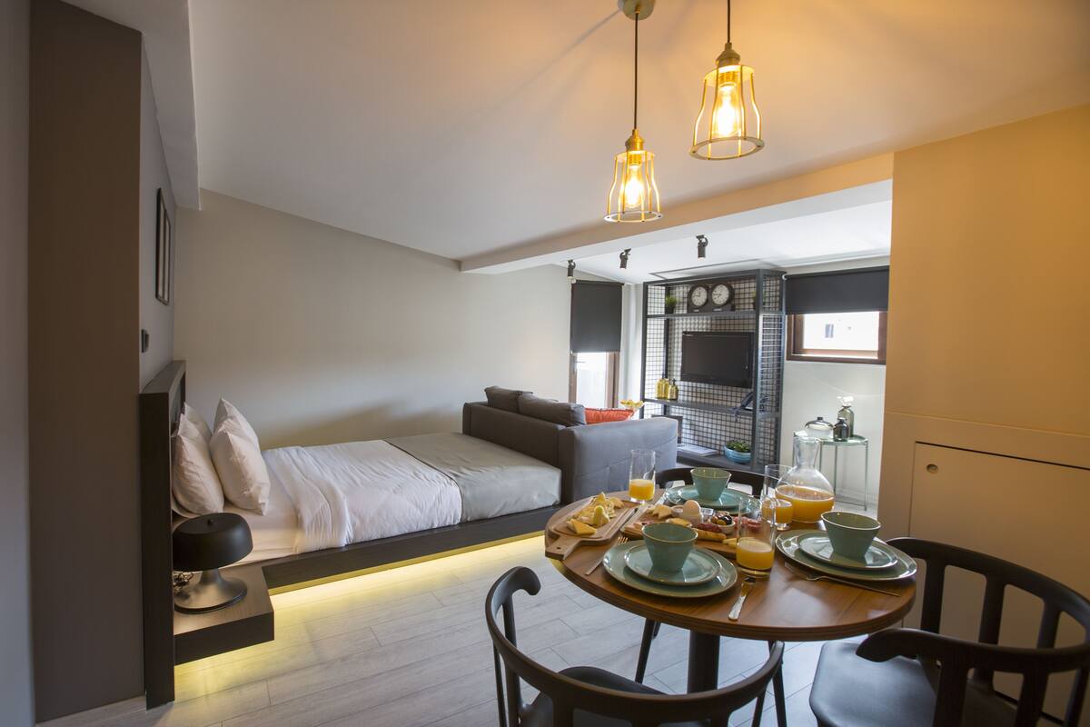 A modern hotel room with a bed, a dining area with breakfast, and a kitchenette.