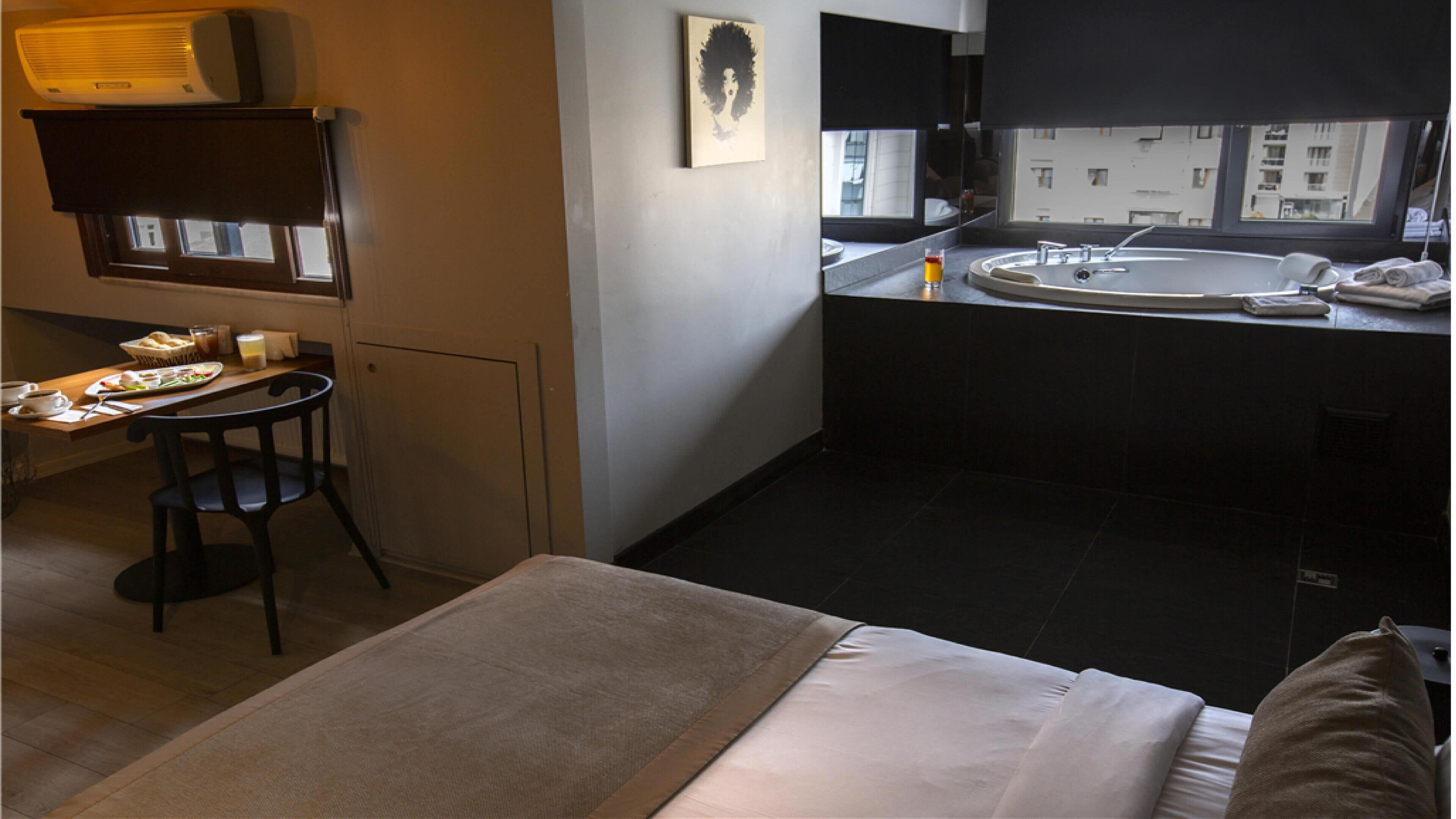 A hotel room with a bed, a small table with breakfast items, a bathroom with a sink and mirror, and a view of buildings through the window.