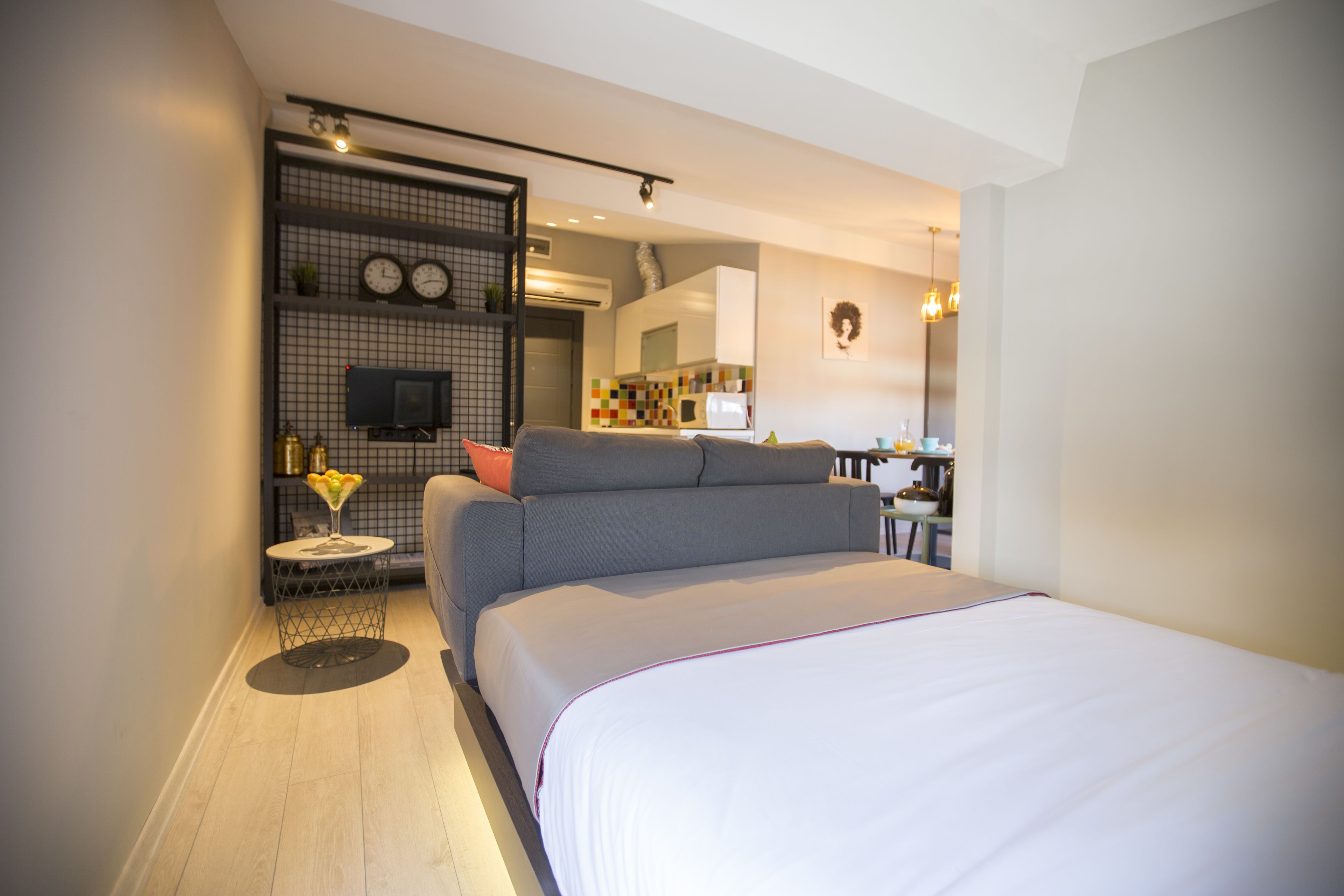 Une chambre moderne avec un grand lit, un canapé gris, une petite table d’appoint avec une lampe et un téléviseur placé sur une étagère murale.