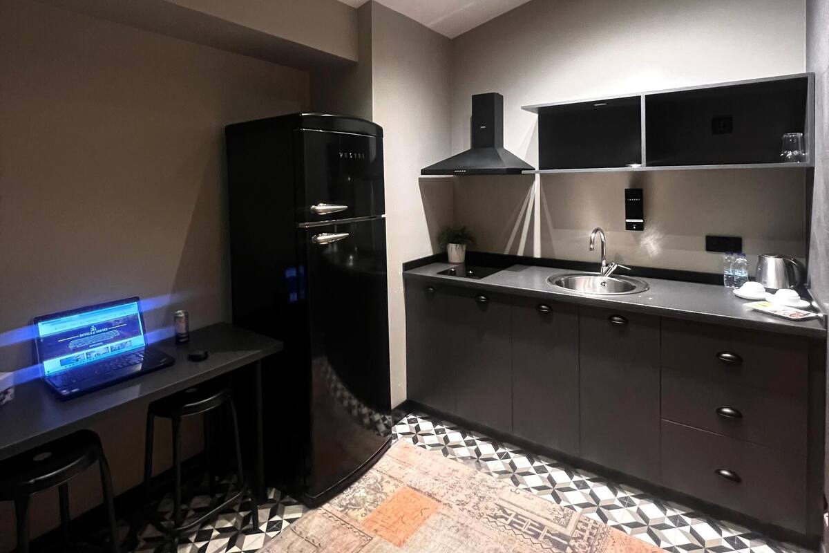 A modern kitchen with a black refrigerator, a built-in stove, and a sink. There is a laptop on the counter and a patterned rug on the floor.