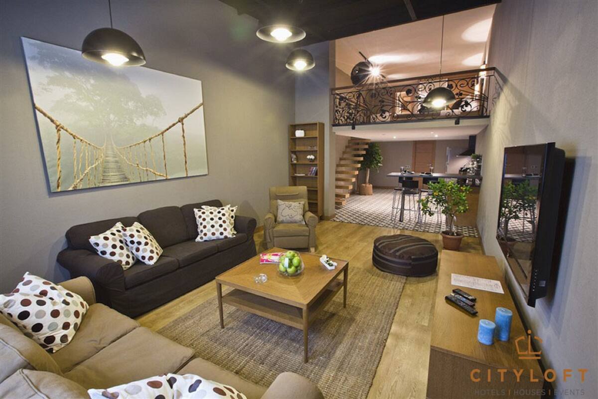 A modern living room with a sofa, coffee table, and a painting of a bridge. There is a staircase leading to a balcony with a railing.