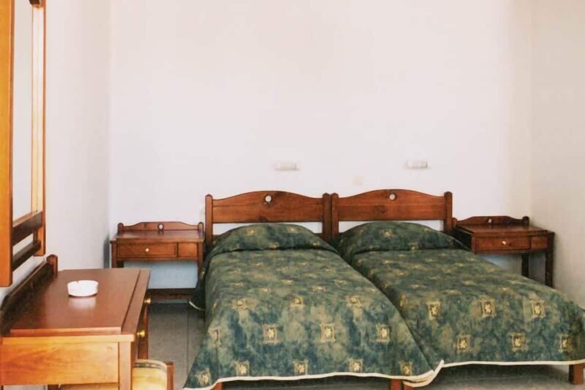 A small, simple room with two beds, a wooden desk, and a chair.