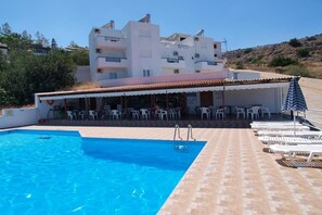 Una piscina con sillas de descanso y mesas, un área de estar techada, y un edificio con varios balcones al fondo.