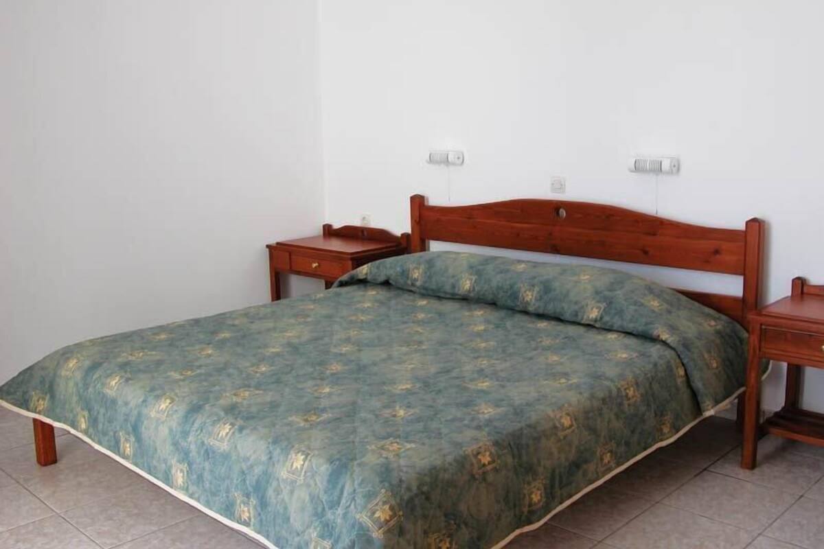 A single-bed room with a wooden headboard, a green patterned bedspread, two bedside tables, and a plain white wall with two electrical outlets.
