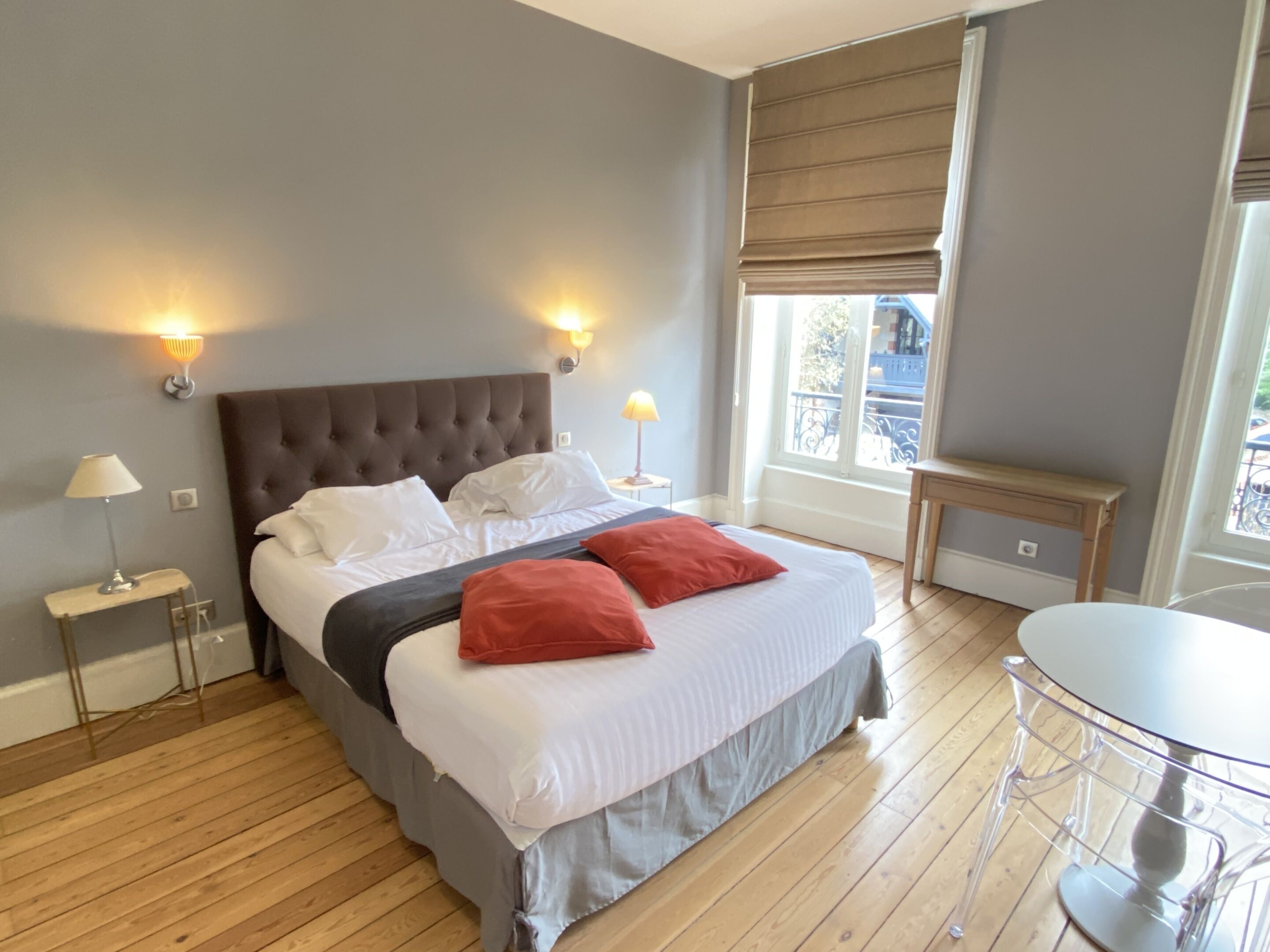 A bedroom with a large bed, two bedside lamps, a wooden floor, a small table, and a window with a view.