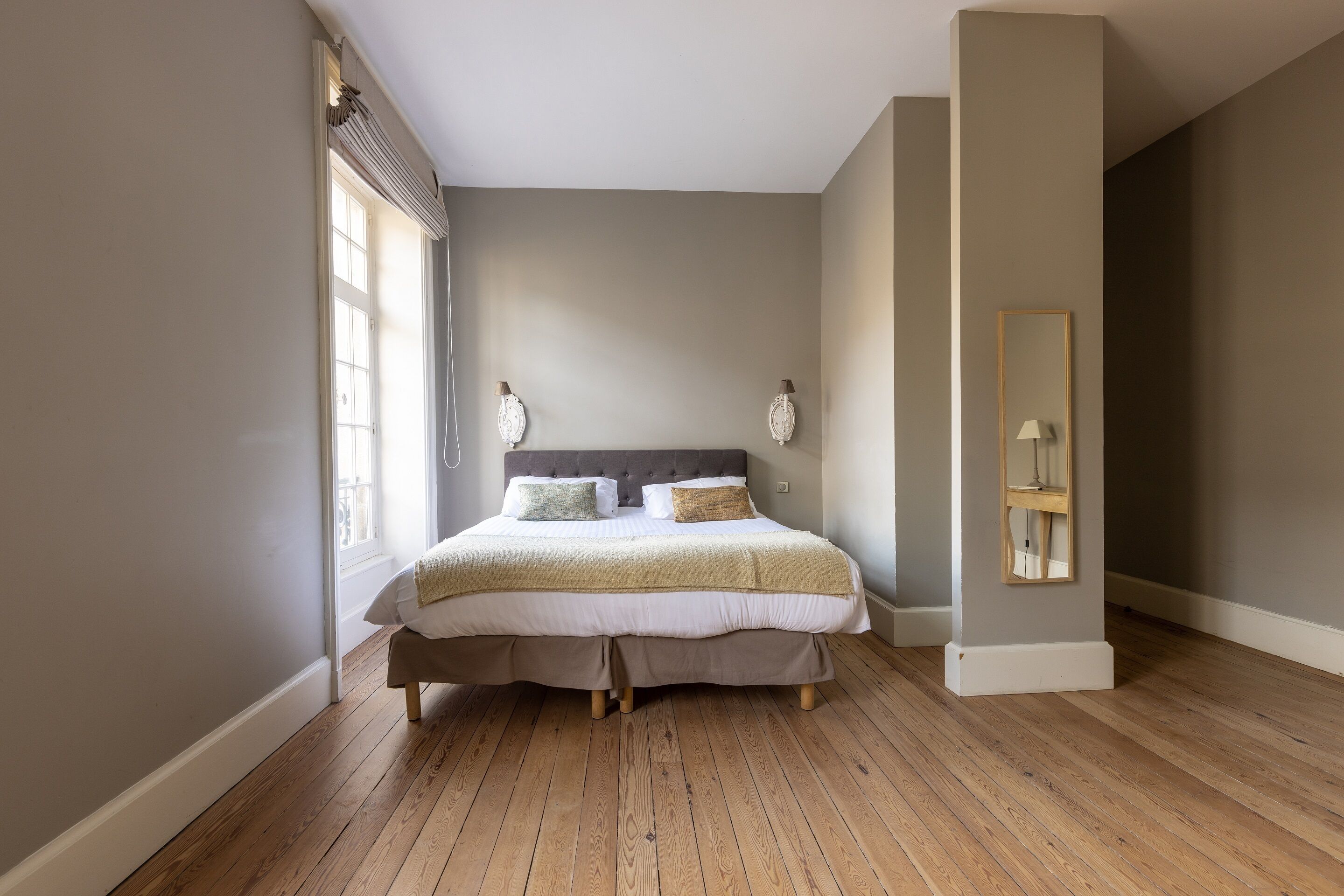 A bedroom with a large bed, a window with blinds, a mirror, and a wooden floor.