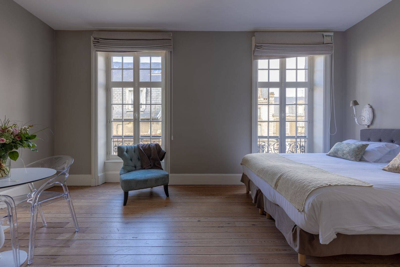 A bedroom with a bed, a chair, a table, and two windows with curtains.